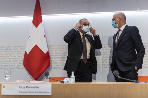 Due uomini in abiti formali con mascherina accanto a bandiera svizzera; sul fondo bianco scritta centro media Palazzo federale