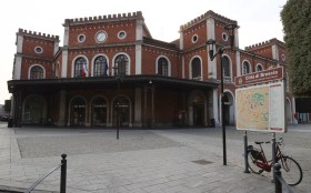 Edificio pubblico im pietre rosse con bandiere italiana ed europea; in primo piano bacheca con mappa di Brescia.
