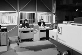 Foto b/n di studio televisivo con 2 giornalisti alla scrivania e pannelli di grafici realizzati a mano
