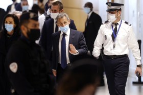 Sarkozy in abiti formali con mascherina attraversa un gruppo di persone/agenti e saluta qualcuno con la mano