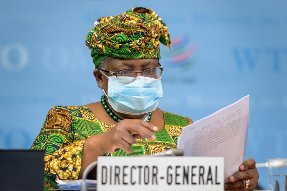 Donna con abito molto colorato e copricapo in tina esamina un dossier; davanti, cartello Director general, dietro logo WTO