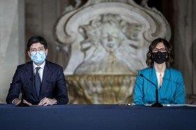 Un uomo (a sx) e una donna (a dx) in abiti formali guardano dritti in camera da un tavolo di conferenza stampa