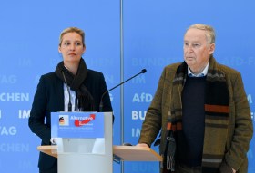 Una donna e un uomo con espressione seria a un pulpito con stemma Alternative für Deutschland, fondo blu
