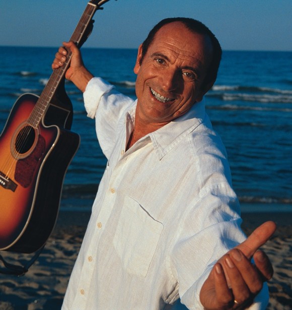 Uomo abbronzato accenna un abbraccio tenendo in mano una chitarra; in riva al mare