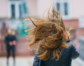 Donna vista di schiena con capelli lunghi, vistosamente sollevati dal vento; ambiente sfocato, forse piazza