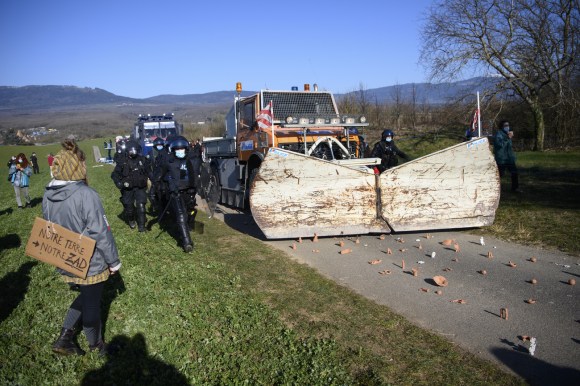 Veicolo stile spazzaneve rimuove oggetti disseminati lungo una strada di collina; accanto, poliziotti in tenuta antisommossa