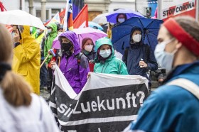 Gruppetto di giovani donne (con impermeabili e mascherine, piove) sfila su una piazzetta con striscioni in tedesco