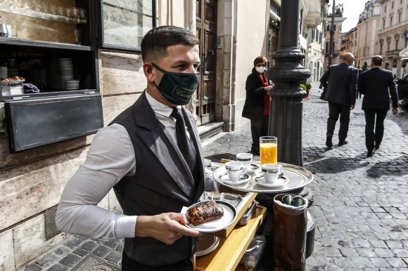 Un cameriere, con la mascherina in volto, porta in mano vassoi con caffè, succhi e brioche.