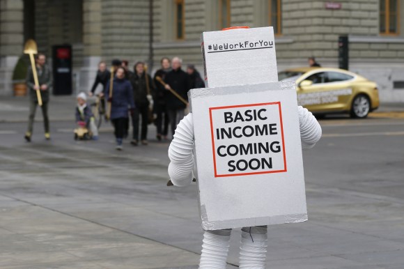 Persona travestita da Robot con alle spalle una scritta: Basic income coming soon