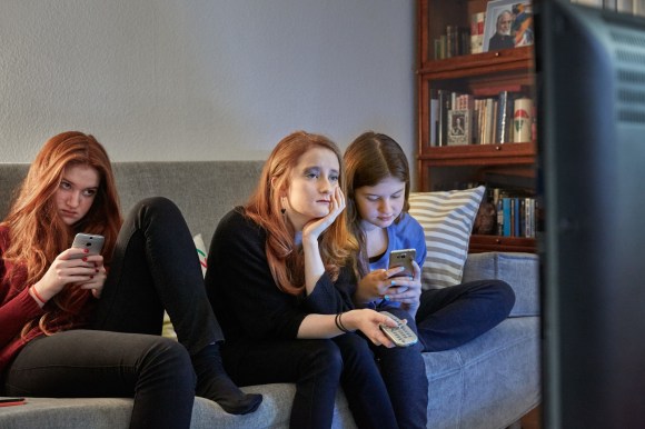 tre ragazze sedute su un divano. Due di loro guardano il telefonino mentre la terza guarda la televisione