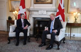 due uomini in giacca e cravatta seduti su due poltrone, dietro ddi loro la bandiera svizzera e quella britannica
