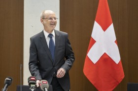 uomo in piedi sorridente, in giacca e cravatta. dietro di lui una bandiera svizzera