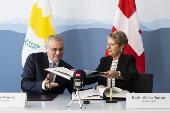 un uomo e una donna si scambiano due cartelle contenenti dei documenti. dietro di loro una bandiera svizzera e una cipriota