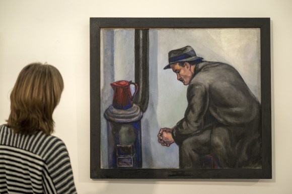 donna fotografata di spalle osserva quadro che ritrae uomo seduto accanto a una stufa sulla quale c è una teiera