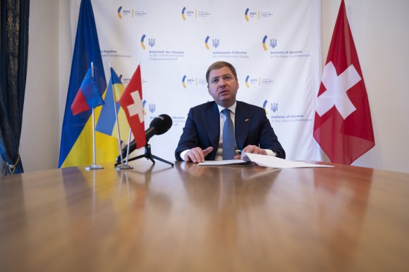 uomo in giacca e cravatta seduto a un tavolo. dietro di lui bandiera svizzera e bandiera ucraina