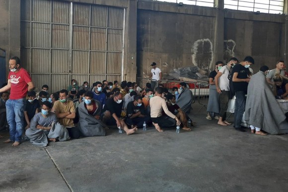 decine di migranti seduti per terra, con coperte sulle spalle e mascherina chirurgica