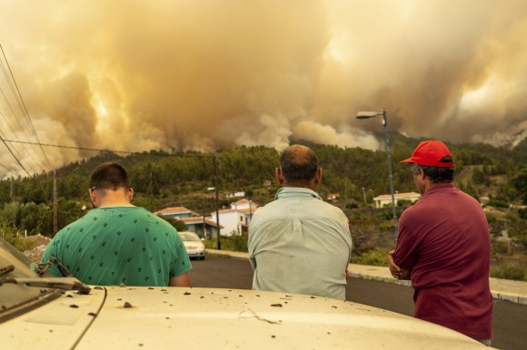 tre uomini appoggiati al cofano di un auto fotografati di spalle osservano un incendio boschivo